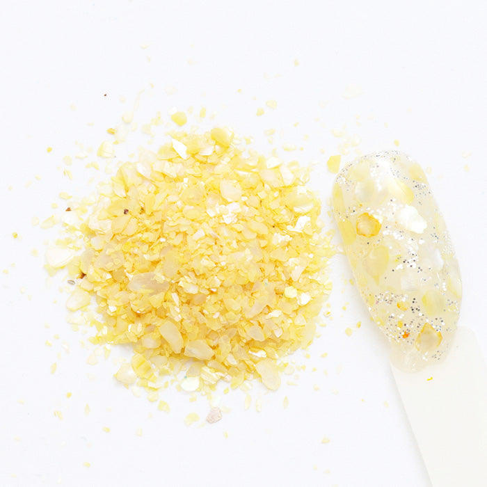 A pile of pale yellow shell flakes used in nail art displayed next to a nail tip on a white background.