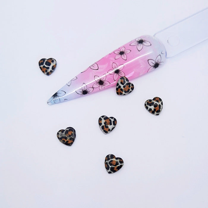 heart shaped nail art charms with a leopard print design placed next to a nail tip on a white background