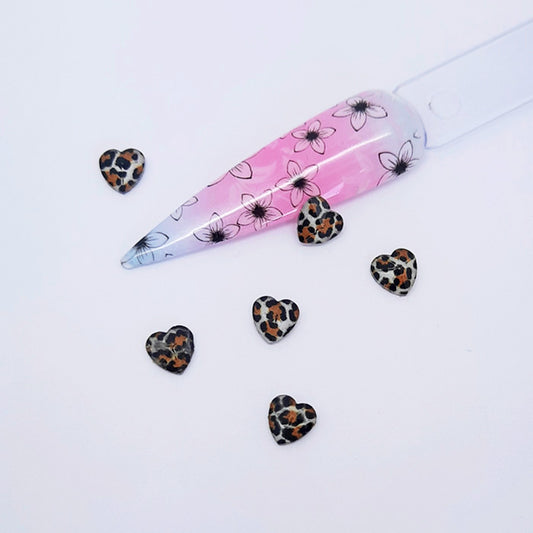 heart shaped nail art charms with a leopard print design placed next to a nail tip on a white background