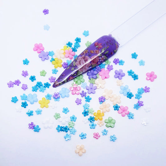 Colourful flower-shaped nail decoration scattered on a light purple background with a decorative purple nail tip.