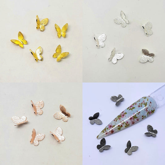 A collection of butterfly-shaped nail charms in various colours including gold, silver, rose gold, and grey, displayed on a white background. 