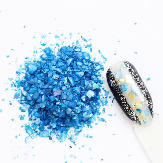 A pile of blue shell flakes used in nail art displayed next to a decorated nail tip on a white background.