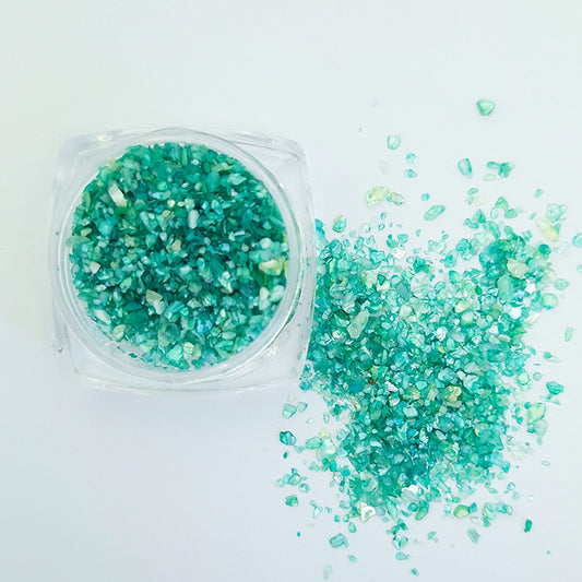 A pile of green shell flakes used in nail art next to a pot of the shell flakes displayed on a white background.