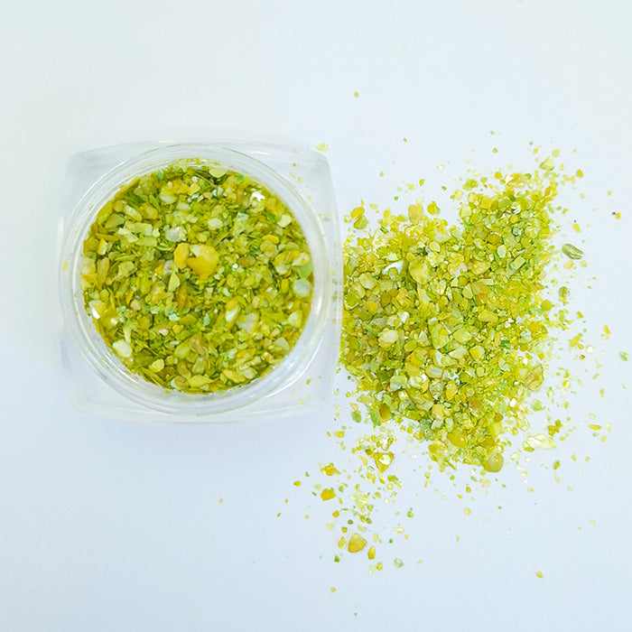 A pile of olive green shell flakes used in nail art spilled from a pot displayed on a white background.