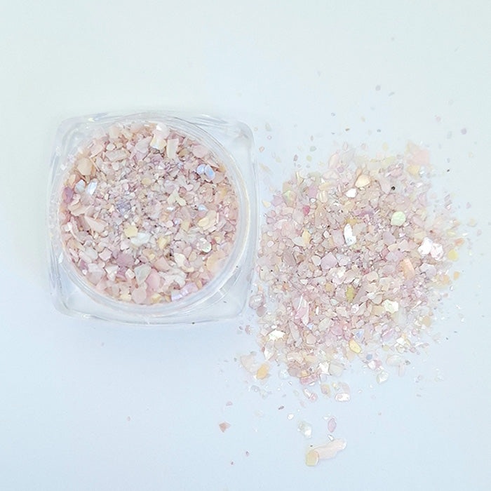 A pile of light pink shell flakes used in nail art spilled from a pot displayed on a white background.